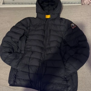 Svart dunjacka Parajumpers med huva - Svart dunjacka från Parajumpers med huva och gul detalj vid halsen. Jackan har quiltad design, dragkedja framtill och en patch på ena ärmen. Perfekt för kalla dagar och har en snygg, sportig look.