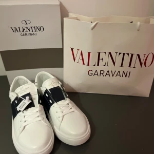 Valentino Garavani Open sneakers vit/blå - Snygga Valentino Garavani Open sneakers i vitt skinn med bred marinblå läderdetalj och silverfärgade nitar på sidorna. Klassisk rund tå, platt sula med mönstrad undersida och vit snörning. Perfekt för dig som gillar exklusiv streetstyle. Skriv om ni vill ha mer bilder eller har några frågor ;)