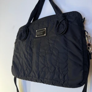 Marc Jacobs datorväska  - Svart väska med handtag och axelrem. Väskan har en silverfärgad metallbricka med texten "MARC BY MARC JACOBS STANDARD SUPPLY TYPE: WORKWEAR". På framsidan finns det stora bokstäver insydda i tyget. Dataväska 