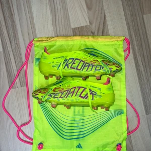 Adidas Predator fotbollsskor  - Säljer ett par Adidas Predator elite fotbollsskor (passar allt mellan strl 40-41) i neon-gul färg med blå och rosa detaljer. Skorna har snörning och en låg profil, designade för maximal kontroll på planen. Snyggt mönster och kommer med matchande väska. Perfekta för dig som vill sticka ut på fotbollsplanen.
