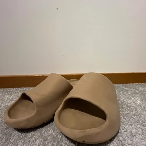 Beige slides med öppen tå - Säljer ett par chunky slides i beige färg med öppen tå och platt sula. De har en minimalistisk design och är tillverkade i syntetmaterial som ger en mjuk och skön känsla. Perfekta för chill dagar eller när du vill ha något enkelt och stilrent på fötterna.