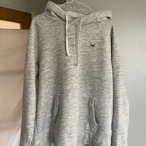 Grå hoodie från Hollister i fleece - Mysig gråmelerad hoodie från Hollister med vit snörning och broderad logga på bröstet. Tröjan har känguruficka, huva och är fodrad med mjuk fleece. Perfekt för kyliga dagar och chill vibes. Tveka inte på att skriva vid funderingar