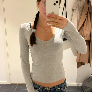 Ljusgrå långärmad croppad topp - Säljer en ljusgrå långärmad topp med v-ringning och croppad passform. Toppen är slim och sitter snyggt mot kroppen. Materialet känns mjukt och stretchigt, troligen bomull eller polyester. Perfekt att styla med jeans eller kjol för en chill look.