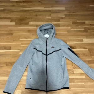 Grå Nike tech fleece med huva - Grå Nike tech fleece med svart logga på bröstet och hel dragkedja framtill. Jackan har huva, svarta detaljer längs bröstet och ärmarna samt praktiska fickor. Perfekt för dig som gillar sportig och clean stil. Storlek S, knappt använd och inga defekter eller sådant!