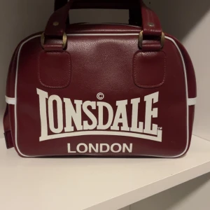 Vinröd handväska - Snygg vinröd handväska från Lonsdale London med vit logga och vita detaljer längs kanterna. Fint skick! Skriv för frågor och funderingar💗vintage! Pris går att diskuteras!!