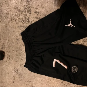 Svarta PSG x Jordan shorts - Säljer ett par svarta PSG x Jordan shorts med elastisk midja. De har en vit Jumpman-logga, nummer 7 och PSG-märke på ena benet. Perfekta för fotboll eller träning, tillverkade i lätt polyester som andas.
