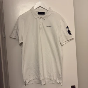Vit Peak Performance piké XL - Klassisk vit pikétröja från Peak Performance i storlek XL. Tröjan har korta ärmar, krage med knappar och svart logga på bröstet samt tryck på ena ärmen. Tillverkad i mjuk bomull som känns skön mot huden.