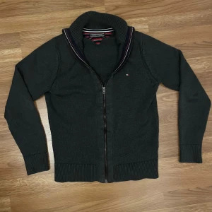 Mörkgrå kofta med dragkedja Tommyy Hilfiger - Mörkgrå stickad kofta från Tommy Hilfiger med ribbad krage och muddar. Dragkedja framtill och liten logga på bröstet. Snygga detaljer med ränder på insidan av kragen i rött, vitt och blått. Perfekt för lager-på-lager under kyliga dagar.