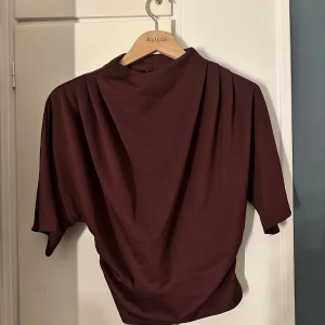 Vinröd topp från Gina Tricot - Säljer en vinröd topp från Gina Tricot i storlek S. Toppen har en tight passform vid midjan med korta ärmar och hög hals. Materialet är mjukt och stretchigt, troligen polyester. Snygga veckade detaljer vid axlarna ger en cool look. 