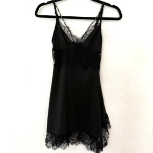 Svart spetsklänning med smala axelband - Supersnygg svart kort klänning med tunna axelband och v-ringning. Klänningen har genomgående spetsdetaljer vid byst och nederkant, vilket ger en extra elegant vibe. Materialet känns mjukt och följsamt, perfekt för dig som gillar en feminin och trendig look.