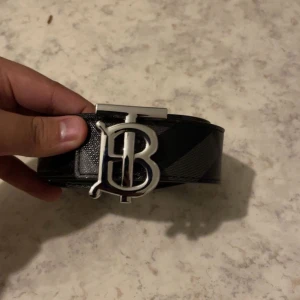 Svart Burberry bälte med silverdetalj - Säljer ett svart Burberry bälte i skinn med klassiskt rutig mönster och en stor silverfärgad spänne med Burberrys ikoniska B-logga. Bältet har en stilren och lyxig vibe som lyfter din outfit. Perfekt för dig som gillar designeraccessoarer.