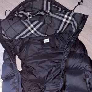 Svart dunjacka från Burberry - Svart dunjacka från Burberry med stor huva och klassiskt rutigt foder i grått och vitt. Jackan har dragkedja, knappar och justerbar nederkant med snörning. Tjock och puffig design som håller dig varm, med tydlig Burberry London England patch på ärmen.