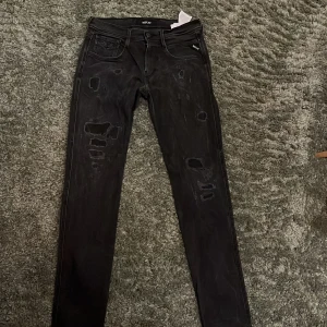 Svarta Replay jeans med slitningar - Säljer ett par svarta Replay jeans i skinny fit med coola slitningar och detaljer på både fram- och baksida. Jeansen har klassisk femficksdesign, dragkedjegylf och är tillverkade i stretchig denim för skön passform.