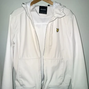 Vit vindjacka från Lyle & Scott - Fräsch vit vindjacka från Lyle & Scott med huva och dragkedja framtill. Jackan har två fickor, ribbad nederkant och det klassiska gula Lyle & Scott-logotypen på bröstet. Perfekt för vår och höst när du vill hålla stilen enkel och clean.dragkejdan har lite problem med att göra sitt jobb bara