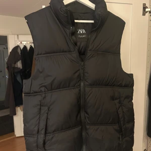 Svart dunväst från Zara, storlek S - Svart dunväst från Zara med hög krage och två sidofickor med dragkedja. Västen har quiltad design och stängs med en kraftig dragkedja framtill. Perfekt för lager-på-lager och har en clean, stilren look som funkar till många outfits.