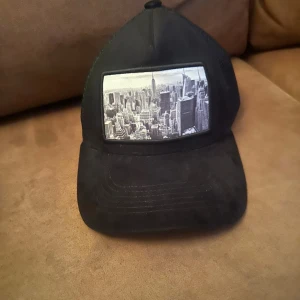 Svart keps med New York-tryck från maggiore  - Svart keps med mesh på sidorna och baksidan för extra ventilation. Framtill finns ett stort tryck med svartvit bild av New Yorks skyline. Justerbar baktill för perfekt passform. Perfekt för dig som gillar urban stil och streetwear.