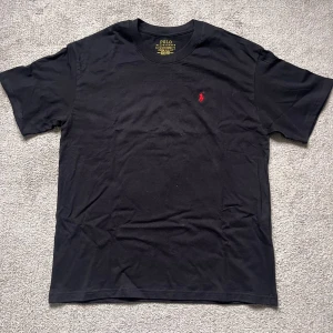 Svart t-shirt från Polo Ralph Lauren - Svart t-shirt från Polo Ralph Lauren med klassisk röd broderad logga på bröstet. T-shirten har rund halsringning och korta ärmar. Tillverkad i mjuk bomull för en skön känsla och enkel stil.