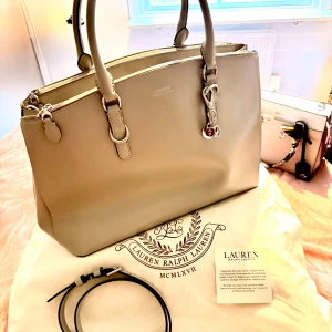Beige handväska från Lauren Ralph Lauren - Snygg beige handväska från Lauren Ralph Lauren i slät skinn med silverfärgade detaljer och dubbla handtag. Väskan har flera fack med dragkedja, innerfack och avtagbar axelrem. Perfekt storlek för allt du behöver och stilren design med logga framtill.