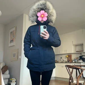 Mörkblå parkas från Helly Hansen S - Parkas från Helly Hansen i mörkblått med röd snörning och stor huva med fluffig fuskpäls. Insidan har röd och blå rutig lining. Jackan har dragkedja, knappar och två stora fickor framtill. 