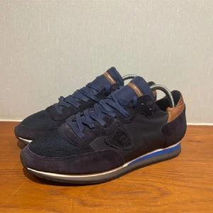 Mörkblå sneakers från Philippe Model - Snygga mörkblå sneakers från Philippe Model med detaljer i brunt skinn vid hälen och på plösen. Ovandelen är en mix av mocka och mesh, med snörning och platt sula. Diskret logga på sidan och blå rand på mellansulan ger en cool touch.