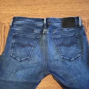 Replay Anbass jeans blå slim fit - Snygga Replay Anbass jeans i blå tvätt med slitningar och klassiska fem fickor. Slim fit-modell med normal midja och raka ben. Jeansen har Replay-logga på myntfickan och detaljerade sömmar på bakfickorna. Perfekt för dig som gillar en modern och stilren look.