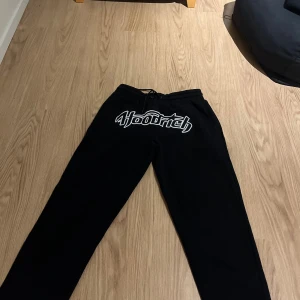 Svarta Hoodrich mjukisbyxor med broderi - Svarta mjukisbyxor från Hoodrich med stort broderat Hoodrich-logo i vitt och svart framtill. Byxorna har elastisk midja med snörning och raka ben. Perfekt för chill eller streetwear-stil.