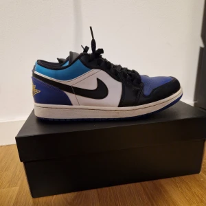 Nike Air Jordan 1 Low blå/svart - Nike Air Jordan 1 (Aqua) Low sneakers med snygg färgblockning i lila, blått, svart och vitt. Skorna har klassisk snörning, rund tå och platt sula. Air Jordan-logga i gult på hälen och svart swoosh på sidan. Perfekt för dig som gillar streetwear och färgglatt. Dem är använda men i fint skick.