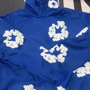 Blå hoodie från Denim Tears med tryck - Säljer en blå hoodie från Denim Tears med unika vita blomliknande tryck över hela tröjan. Hoodien har huva, långa ärmar och känguruficka framtill. Perfekt statement-plagg för dig som vill sticka ut med cool design.