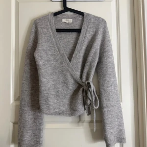 Grå omlottkofta från Gina Tricot - Mysig grå omlottkofta från Gina Tricot i ullblandning. Koftan har långa ärmar, ribbade muddar och knytband i sidan för en snygg passform. Perfekt för lager-på-lager och chill dagar.