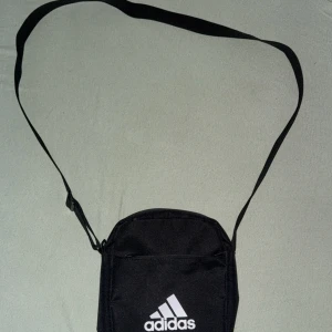 Svart axelväska från Adidas - Svart axelväska från Adidas med justerbar rem och klassisk vit logga framtill. Väskan har flera fack med dragkedja och är tillverkad i slitstark polyester. Perfekt storlek för att bära med sig det viktigaste och enkel att matcha med olika outfits.