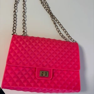 Rosa axelväska med kedja - Snygg rosa axelväska med coolt geometriskt mönster och silverfärgad kedja. Väskan har ett vridlås i metall och är gjord i syntetmaterial. Perfekt för dig som vill sticka ut med en färgglad accessoar. Rymmer mobil, plånbok och småsaker.