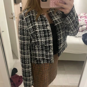 Svartvit rutig kavaj med knappar - Säljer en svartvit kavaj med rutigt mönster och snygga silvriga knappar framtill. Jackan är croppad och har lång ärm, perfekt för att ge din outfit en chic vibe. Materialet är strukturerat och känns lyxigt, med klassisk krage och stilren passform. S/M