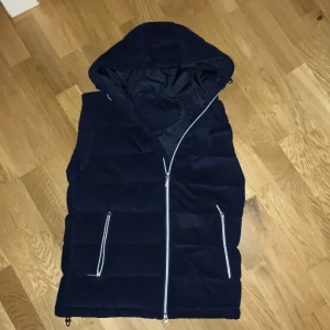 Mörkblå dunväst från Vendiera - Snygg mörkblå dunväst från Vendiera med huva och silverfärgade dragkedjor. Västen är quiltad och har två praktiska fickor med dragkedja. Yttermaterialet är manchester vilket ger en cool och unik look. Perfekt för lager-på-lager under kyliga dagar.