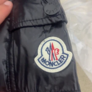 Svart Moncler pufferjacka med huva - Svart pufferjacka från Moncler med glansig finish och huva. Jackan har dragkedja framtill, fickor med dragkedja och klassisk Moncler-logga på ärmen. Insidan har en cool serietidningsetikett och jackan är certifierad med DIST för dunets kvalitet.