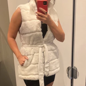 Vit pälsväst med bälte - Säljer en fluffig vit pälsväst med bred krage och snyggt bälte i midjan. Västen har fickor på sidorna och är i en loose passform. Perfekt för att addera lite lyx till din outfit och hålla dig varm under kyliga dagar.