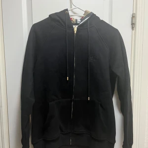 Svart hoodie från Burberry med dragkedja - Svart hoodie från Burberry med guldfärgade detaljer på snörningen och dragkedja. Klassisk huva, stora fickor framtill och Burberry-logga broderad på bröstet. Insidan av huvan har det ikoniska rutmönstret. Perfekt för chill dagar.