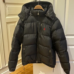 Polo Ralph Lauren vinterjacka - Svart pufferjacka från Polo Ralph Lauren med huva, dragkedja och flera fickor med zip. Jackan har det klassiska röda Polo-logot på bröstet och justerbar nederkant med snörning. Perfekt för kalla dagar och har en schysst, lite boxig passform. Användes under förra vintern!