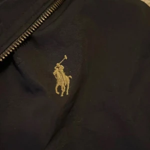 Svart vindjacka från Polo Ralph Lauren - Svart vindjacka från Polo Ralph Lauren med klassisk broderad logga på bröstet. Jackan har huva, dragkedja framtill och justerbara detaljer vid ärmslut och midja. Perfekt för blåsiga dagar och har en stilren look som funkar till det mesta.