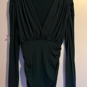 Mörkgrön omlottklänning från SHEIN - Supersnygg mörkgrön kort klänning från SHEIN med omlott och djup v-ringning. Klänningen har långa ärmar, rynkade detaljer vid axlar och midja samt figurnära passform. Perfekt för dig som vill sticka ut med en elegant men enkel look.