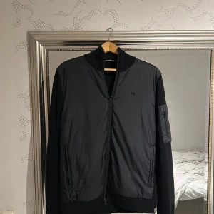 Svart bomberjacka från J.Lindeberg - Svart cardigan  från J.Lindeberg med stilren design och diskret logga på bröstet. Jackan har ribbad krage och muddar, dragkedja framtill samt fickor med dragkedja. Materialmix med polyester på framsidan och stickad bomull på ärmar och rygg. Perfekt för dig som gillar clean look.