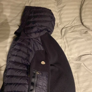 Mörkblå Moncler pufferjacka med huva - Säljer en mörkblå pufferjacka från Moncler med stickade paneler och quiltad huva. Jackan har dragkedja framtill och en ficka med dragkedja på ärmen. Ikonisk Moncler-logga på armen och snygg, modern look som passar till streetwear.