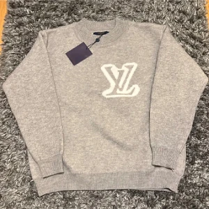 Grå stickad Louis Vuitton tröja  - Exklusiv grå stickad tröja från Louis Vuitton med stort vitt LV-logo framtill. Rund hals och ribbade muddar vid ärmslut och nederkant. Perfekt för dig som gillar lyxiga och stilrena plagg med statement-detaljer.