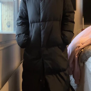 Svart lång pufferjacka med hög krage - Säljer en svart lång pufferjacka med hög krage från Kappahl. dragkedja samt knappar framtill. Jackan har en clean look, stora fickor och är perfekt för kalla dagar, även knappar längst ner vid kanterna som går att öppna, Snygg och enkel att styla till vintern. Använt fåtalgåmger, ny pris 1799kr
