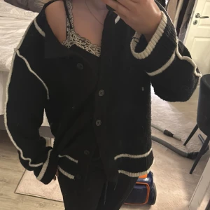 Svart kofta med vita detaljer - Säljer en svart oversized kofta med vita ränder längs ärmar, muddar och nederkant. Koftan har knappar framtill och är stickad i ett mjukt material. I storlek M