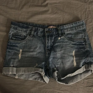 Blå jeansshorts från Miss Milla - Säljer ett par blå jeansshorts från Miss Milla med slitna detaljer och upprullade benslut. Klassisk femficksmodell med bälteshällor och knappgylf. Perfekta för varma dagar och en avslappnad look.
