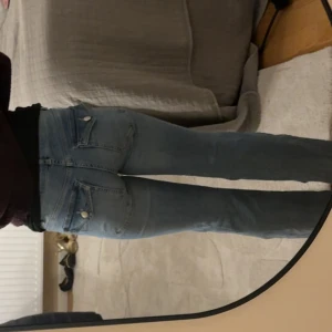 Bootcut ljusblå jeans med fickor - Säljer ett par ljusblå bootcut jeans med klassiska fem fickor och knappstängning fram. Jeansen har en rak passform över låren och lätt utsvängda ben. Materialet är jeans och de har en snygg, enkel look som passar till sneakers eller boots.