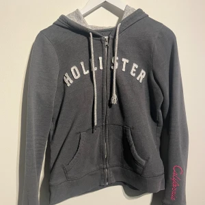 Hollister Grå Hoodie - Grå Hollister hoodie med dragkedja och fickor fram. Applikation med märkets namn framtill och broderi på ena ärmen.  Skick: Gott skick men väl använd. Normalt. bruksslitage. På en av luv snörerna så har den lilla plastbiten fallit av.  Storlek: M men passar även S.  Kolla gärna in mina andra annonser.   Skriv vid frågor eller ifall du vill ha fler bilder. 