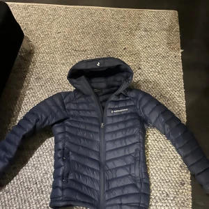 Mörkblå dunjacka Peak Performance S - Snygg mörkblå dunjacka från Peak Performance i storlek S. Jackan har huva, quiltad design och dragkedja framtill. Perfekt för kalla dagar och har en clean look med logga på bröstet. Passar dig som gillar sportig och stilren vibe.