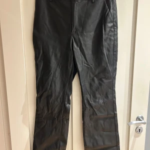 Svarta skinnbyxor från Gina Tricot - Snygga svarta byxor i skinnimitation från Gina Tricot. Klassisk rak passform med hällor för bälte, sidfickor fram och dekorativa fickor bak. Perfekta för en edgy look och lätta att matcha med allt.