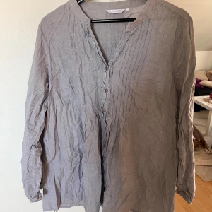 Grå henley från Cappuccini, strl 46 - Säljer en grå henley-skjorta från Cappuccini i storlek 46. Skjortan har lång ärm, knappar framtill och snygga rynkade detaljer längs knappslån. Materialet är lätt och känns som bomull.